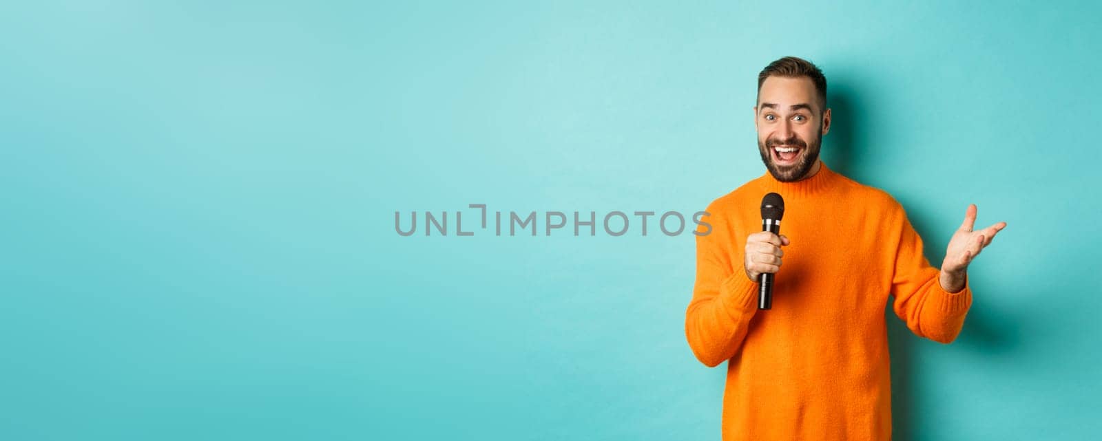 Handsome adult man perform song, singing into microphone, standing against turquoise background.