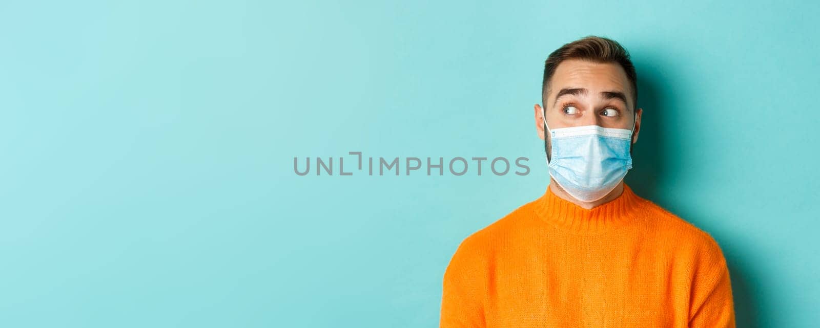 Covid-19, social distancing and quarantine concept. Close-up of young male model in medical mask looking left curious, staring at promo copy space, light blue background.