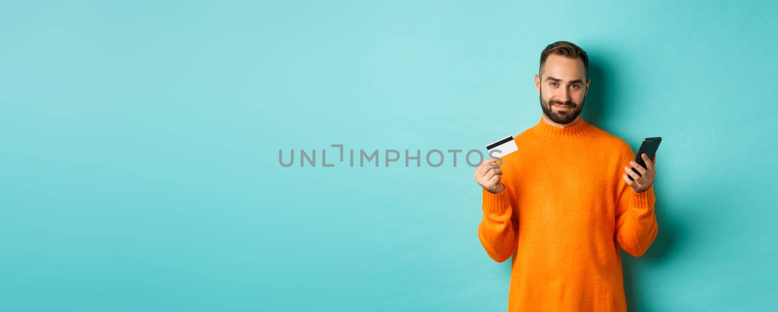 Online shopping. Handsome caucasian man in orange sweater, using credit card and mobile phone, standing over light blue background.