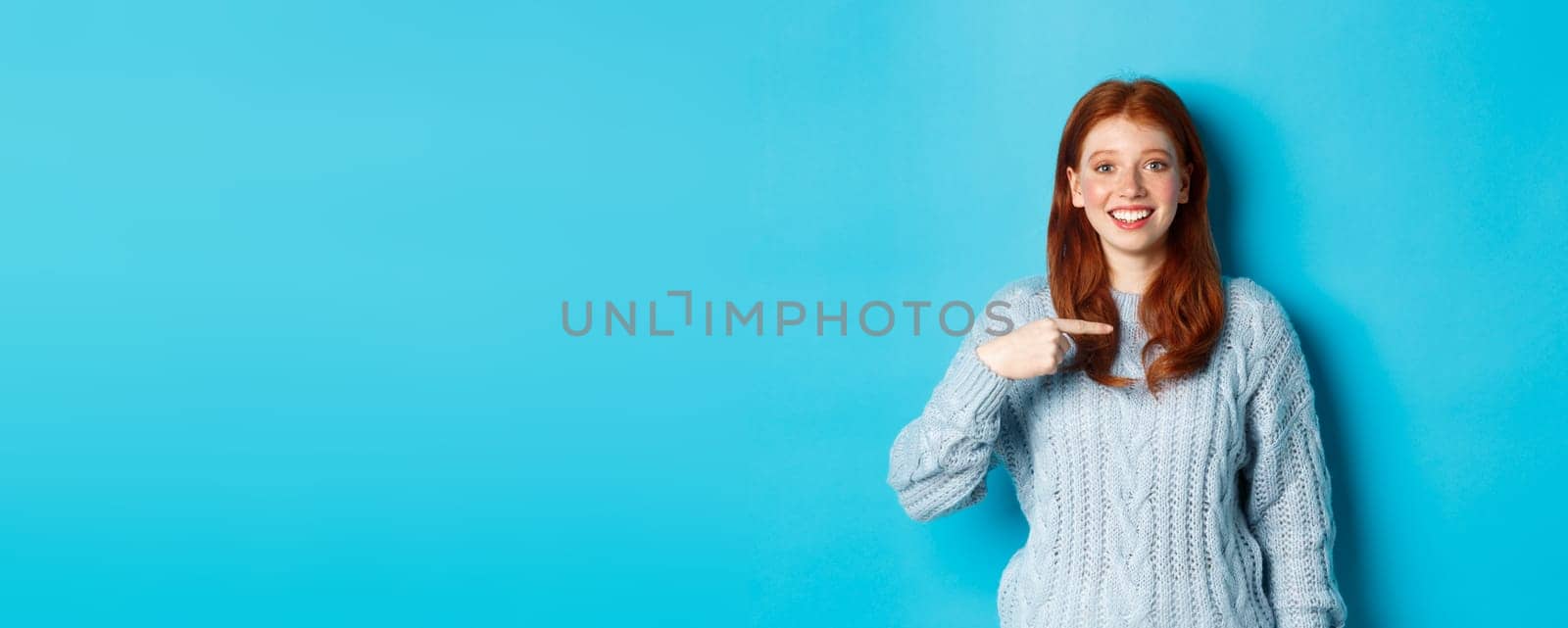 Beautiful redhead girl pointing at herself and smiling happy, being chosen, standing in sweater against blue background.