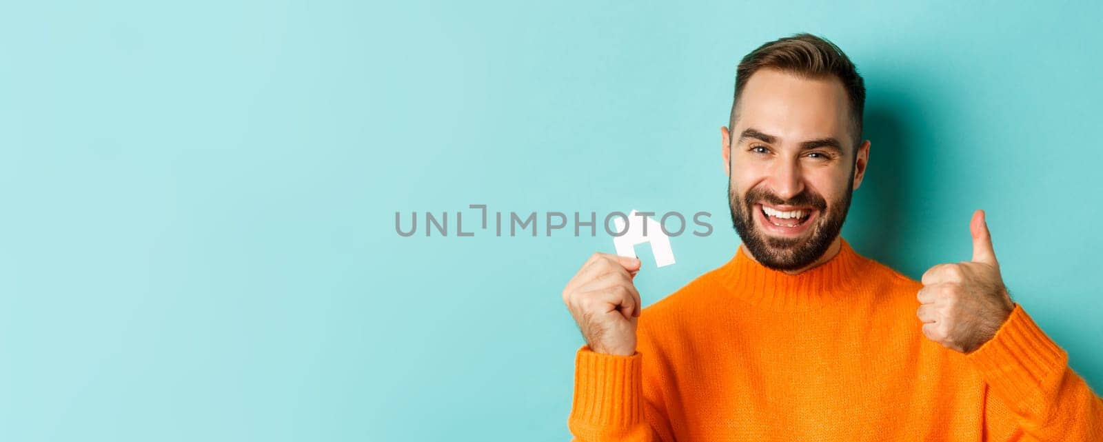 Real estate. Close-up of satisfied man showing thumb-up and small paper house maket, smiling pleased, standing over light blue background.