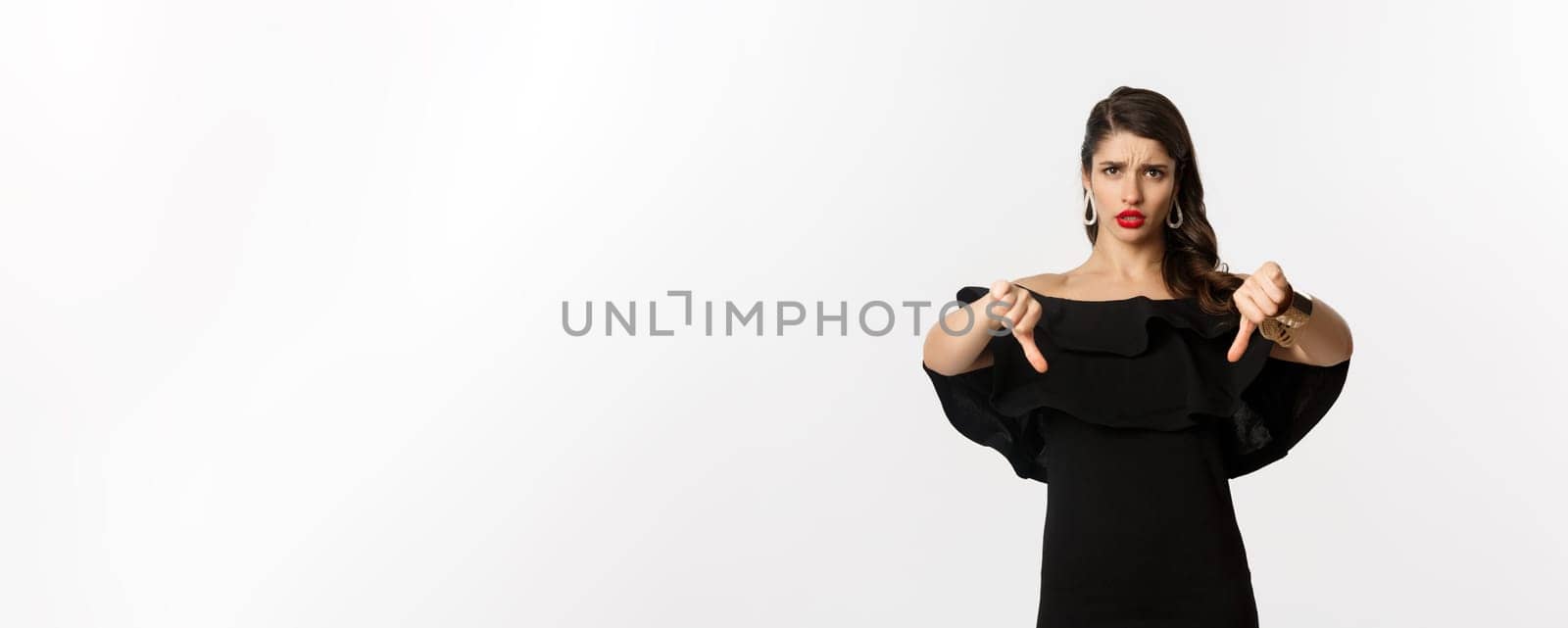 Fashion and beauty. Disappointed and upset woman in black dress, showing thumbs down, dislike something bad, judging over white background.