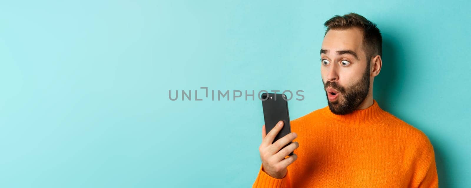 Close-up of caucasian man staring at phone screen with surprised face, wearing orange sweater, light blue background.
