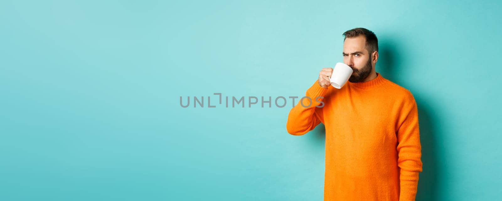 Image of guy drinking coffee and looking suspicious at camera, stare at something strange, standing in orange sweater over turquoise background.