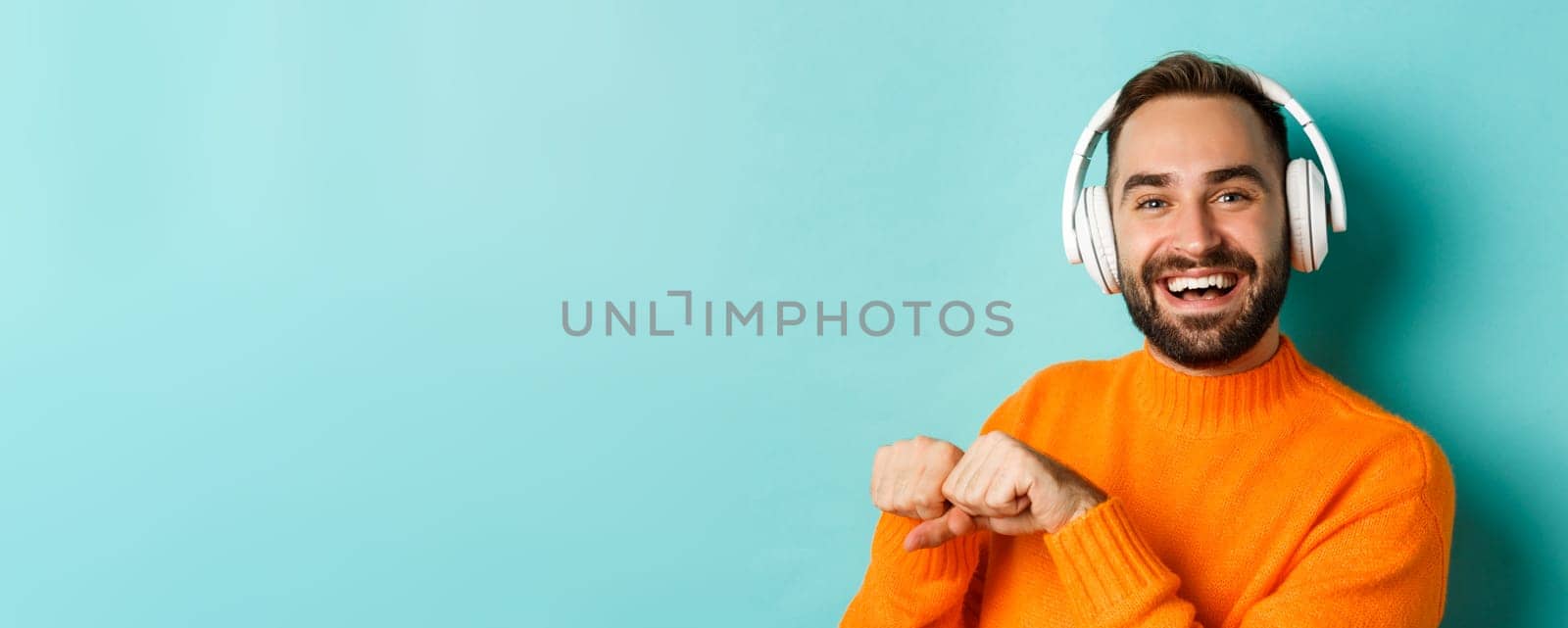 Close-up of handsome modern man listening music in headphones, standing in orange sweater over turquoise background by Benzoix