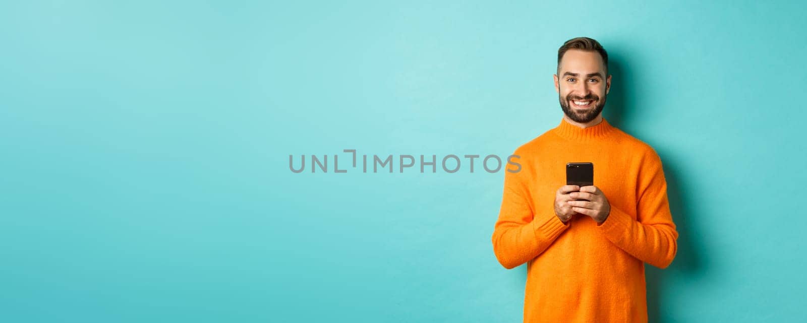 Happy handsome man writing message on mobile phone, holding smartphone and smiling, standing against turquoise background.