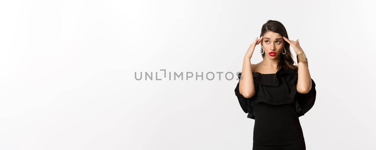 Fashion and beauty. Annoyed and tired woman in black dress, touching head and roll eyes bothered, standing distressed against white background.