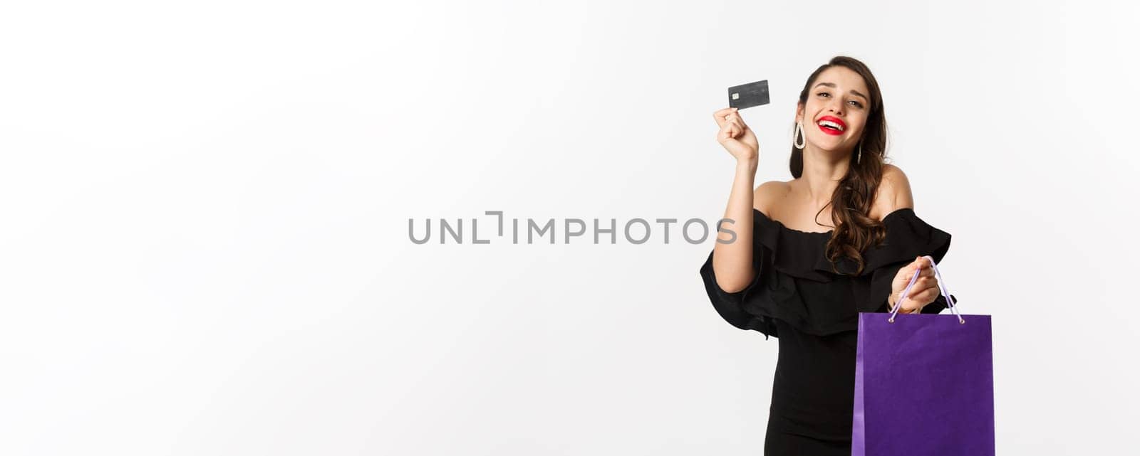 Stylish young woman in black dress going shopping, holding bag and credit card, smiling pleased, standing over white background.