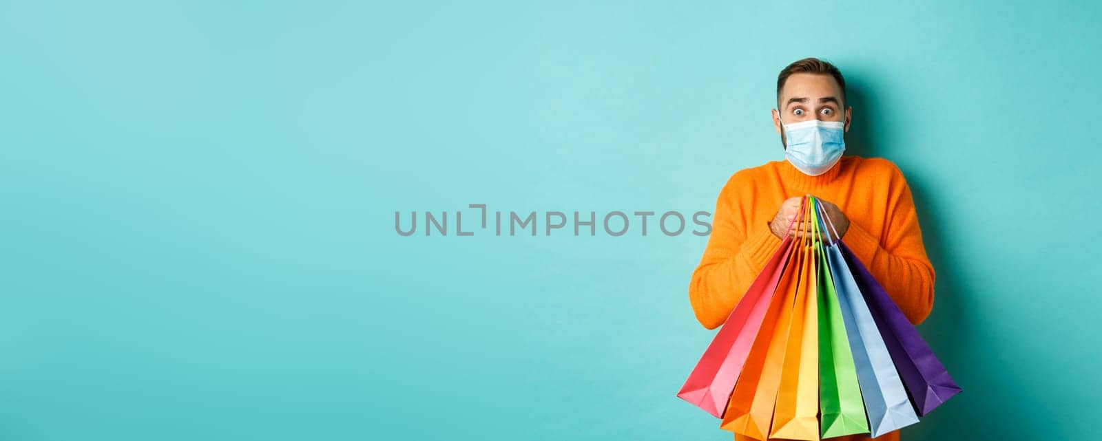 Covid-19, pandemic and lifestyle concept. Excited man in face mask showing shopping bags and rejoicing from discounts, standing over turquoise background.