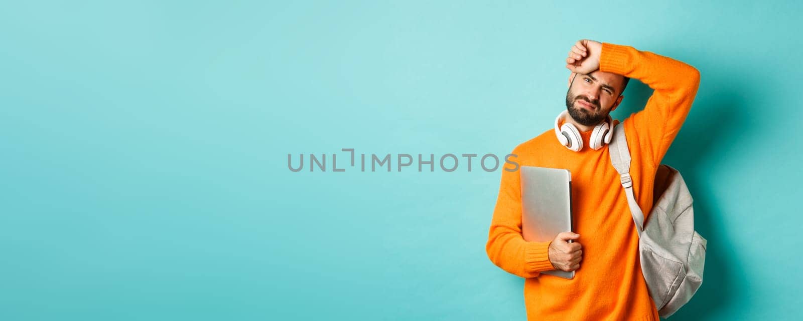 Tired male student wiping sweat off forehead, holding laptop and backpack, standing in orange sweater by turquoise background.