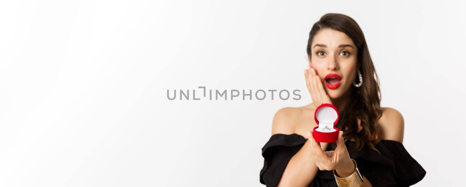 Beautiful woman in black dress making proposal, showing engagement ring and looking excited, standing over white background.