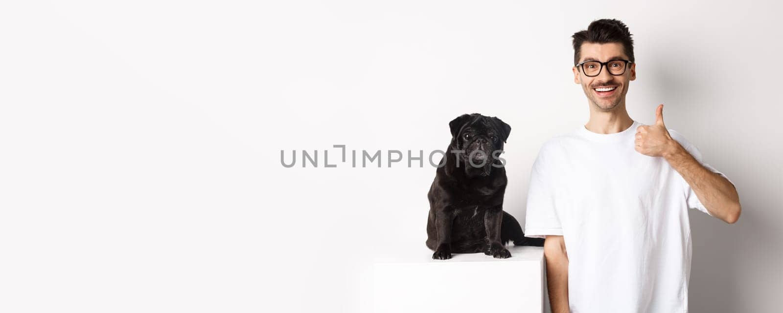 Image of dog owner and cute black pug looking at camera, man showing thumb-up in approval, recommending something, standing over white background.