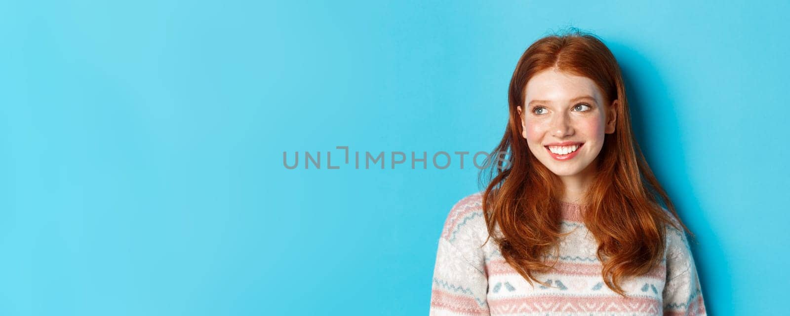Close-up of beautiful redhead female model in winter sweater, smiling and looking left at copy space, standing over blue background by Benzoix
