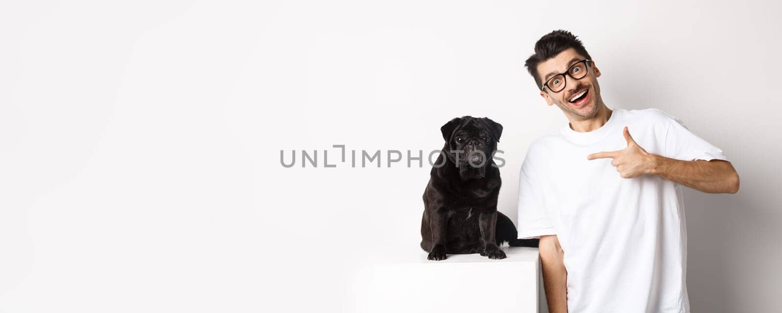 Happy young man showing his cute dog, pointing finger at black pug and smiling, standing over white background by Benzoix