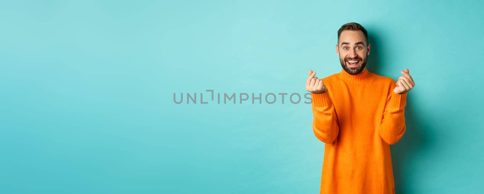 Smiling handsome man showing hearts and looking happy, lucky gesture, standing in orange sweater over light blue background.