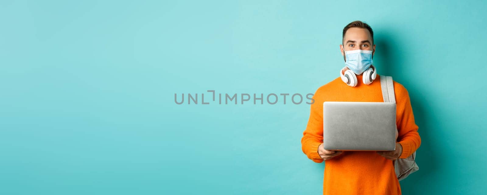 Young man in face mask using laptop and staring happy at camera, standing over light blue background by Benzoix