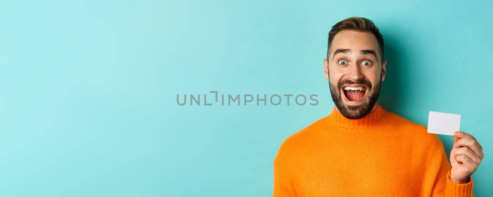 Close-up of excited caucasian man showing his credit card, smiling and staring amazed, standing in orange sweater against turquoise background.
