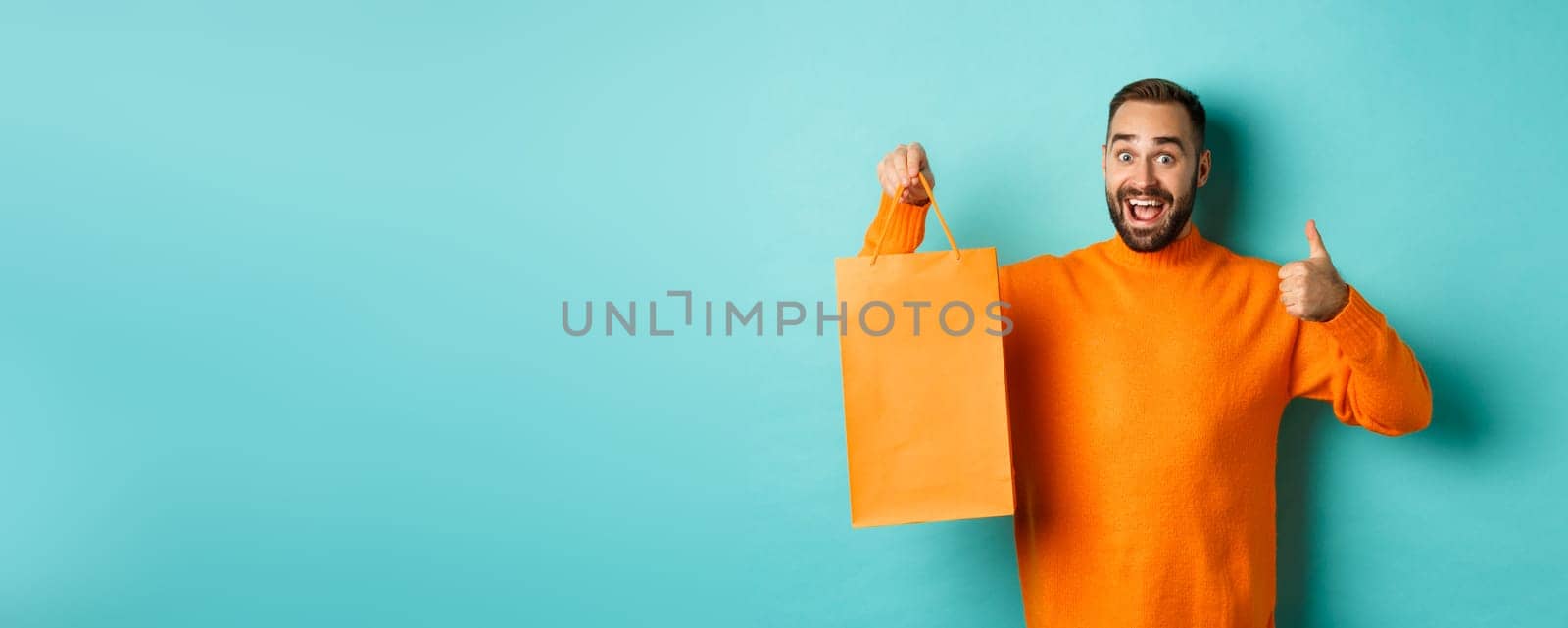 Happy man holding orange shopping bag and rejoicing from discount and celebrating, showing thumbs up and recommending, standing over blue background by Benzoix