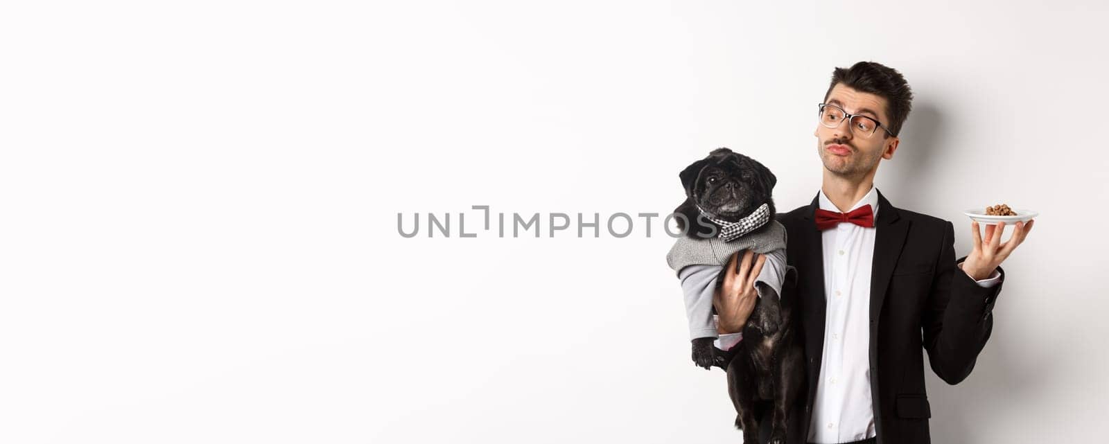 Handsome young dog owner in fancy suit holding cute black pug and plate with animal food, standing over white background.