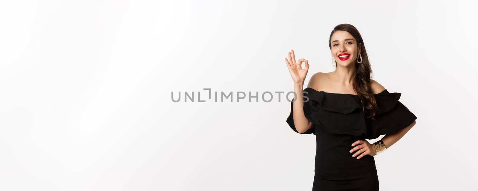 Fashion and beauty. Satisfied good-looking woman with red lipstick, black dress, showing okay sign in approval, like and agree, standing over white background.
