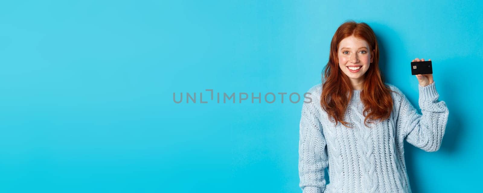 Cute redhead girl in sweater showing credit card, smiling at camera, standing over blue background by Benzoix