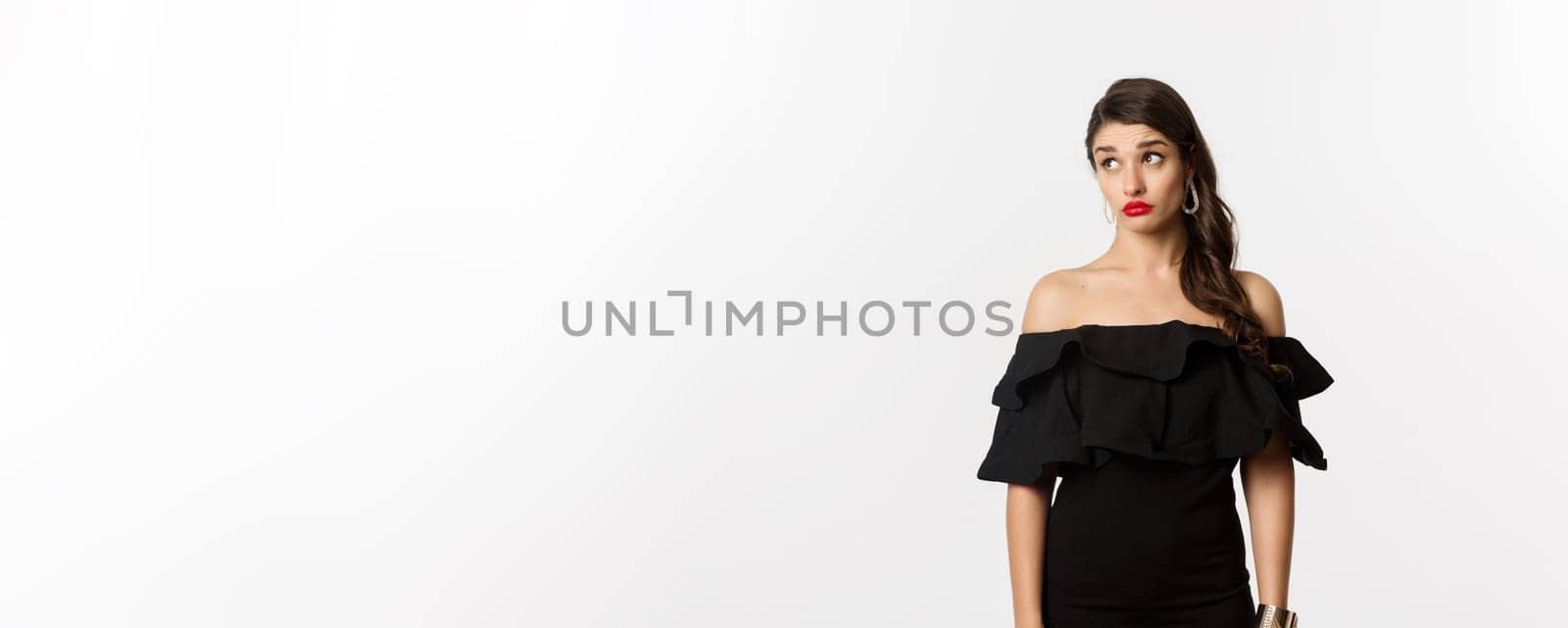 Fashion and beauty. Confused attractive woman in black dress, looking away with puzzled and shocked face, standing over white background.