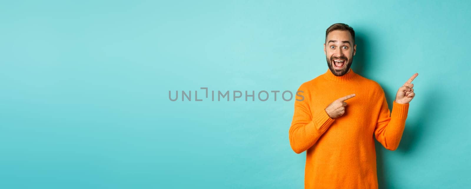 Excited man making an announcement, pointing fingers right your logo, standing over turquoise background.