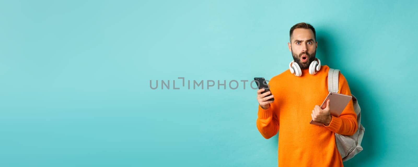 Handsome man student with headphones and backpack, holding digital tablet and smartphone, staring confused at camera, standing against turquoise background.