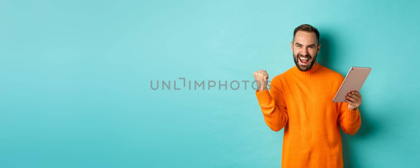 Cheerful winning man holding digital tablet, rejoicing and celebrating victory in game, making fist pump gesture, standing over light blue background.