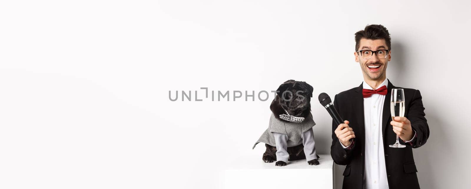 Happy guy raising glass of champagne, giving microphone to dog in costume, celebrating and enjoying party, standing over white background by Benzoix