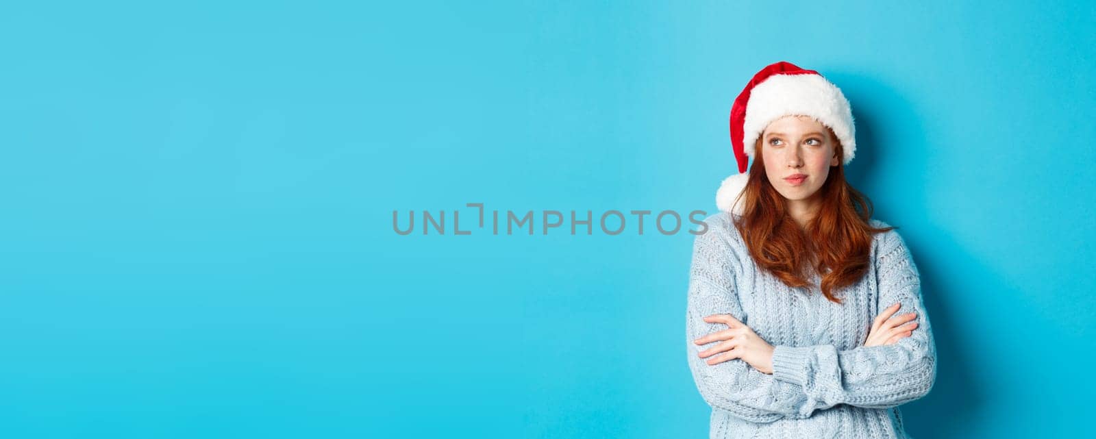 Winter holidays and Christmas Eve concept. Thoughtful redhead woman in Santa hat and sweater, looking left and pondering, making xmas plans, standing over blue background by Benzoix
