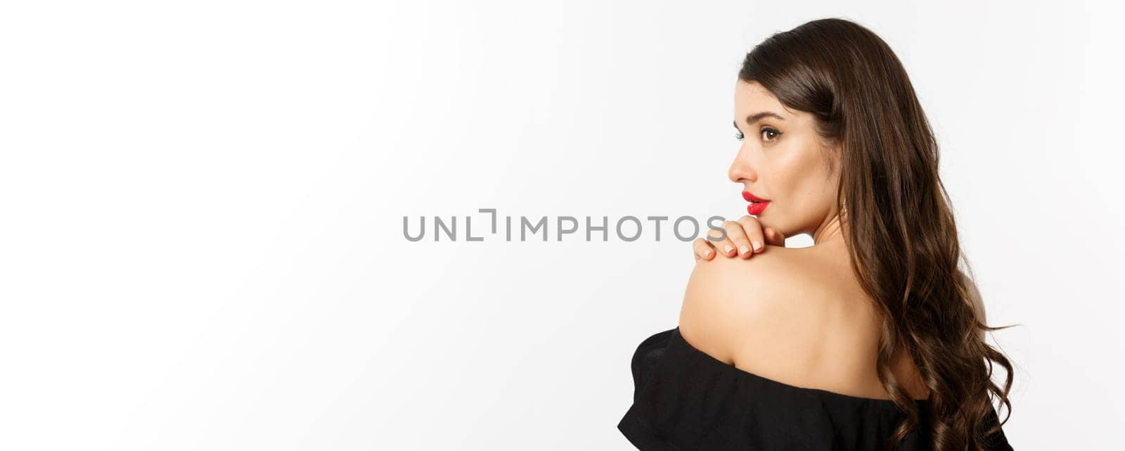 Fashion and beauty concept. Elegant woman leaning on shoulder and gazing aside with sensual piercing eyes, wearing makeup and red lipstick, standing over white background.