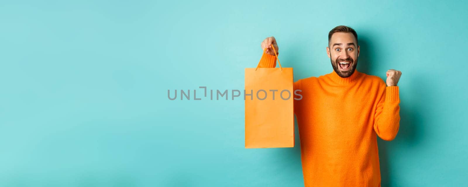 Happy man holding orange shopping bag and rejoicing, got discount and celebrating, standing over turquoise background.