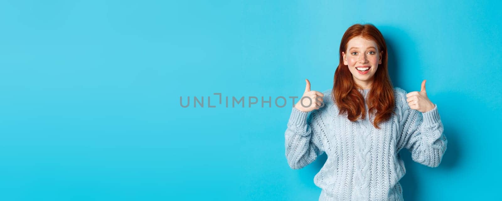 Cheerful teen girl with red hair, showing thumbs up in approval, like and praise gesture, standing over blue background.