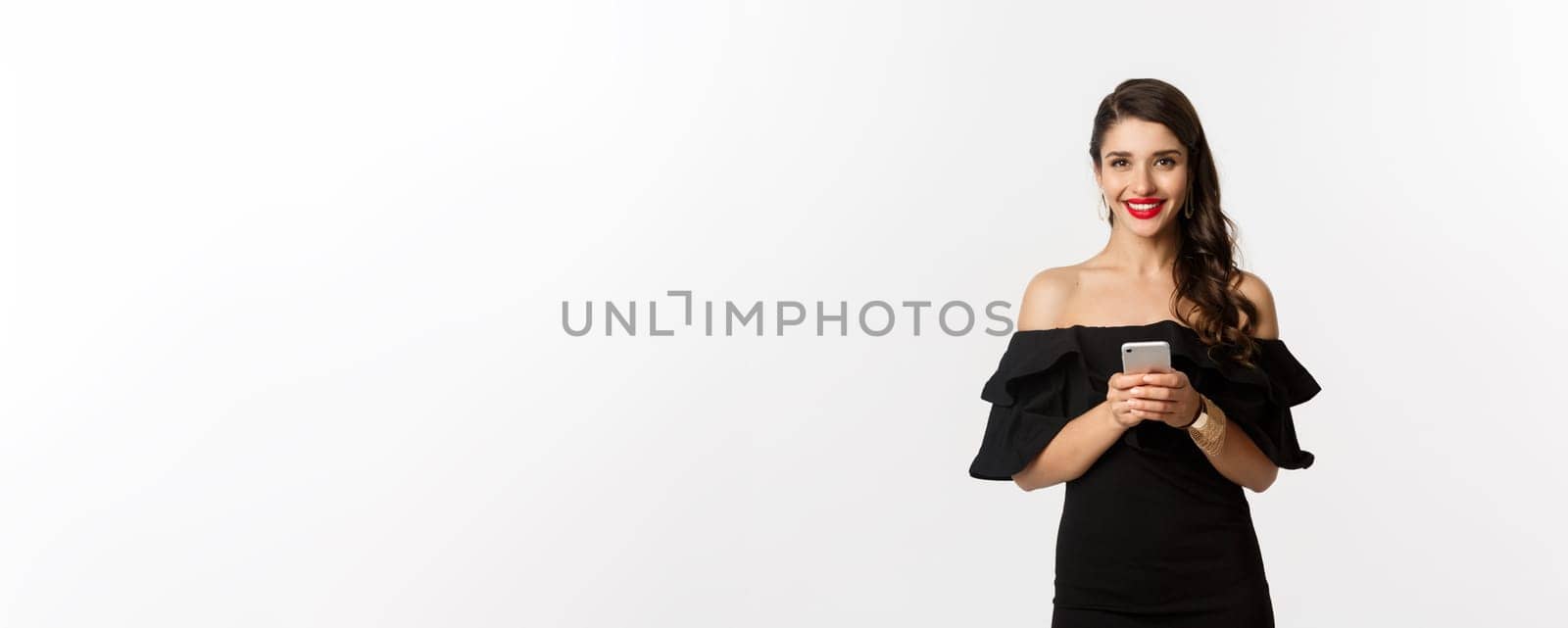 Online shopping concept. Attractive young woman in black dress, reading text message, using mobile phone and smiling, standing over white background by Benzoix