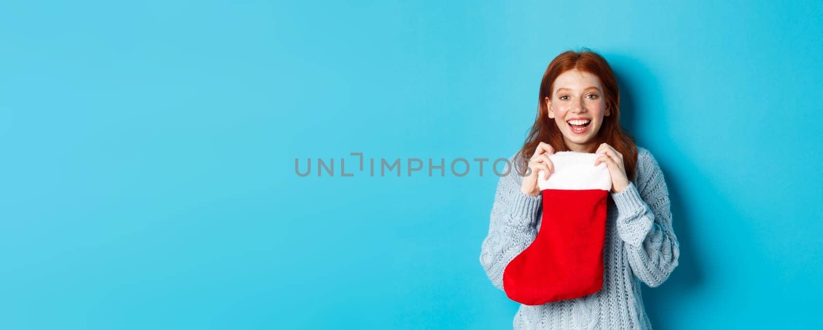 Winter holidays and gifts concept. Happy teenage redhead girl receiving xmas gift, open christmas stocking and smiling amazed, standing over blue background.