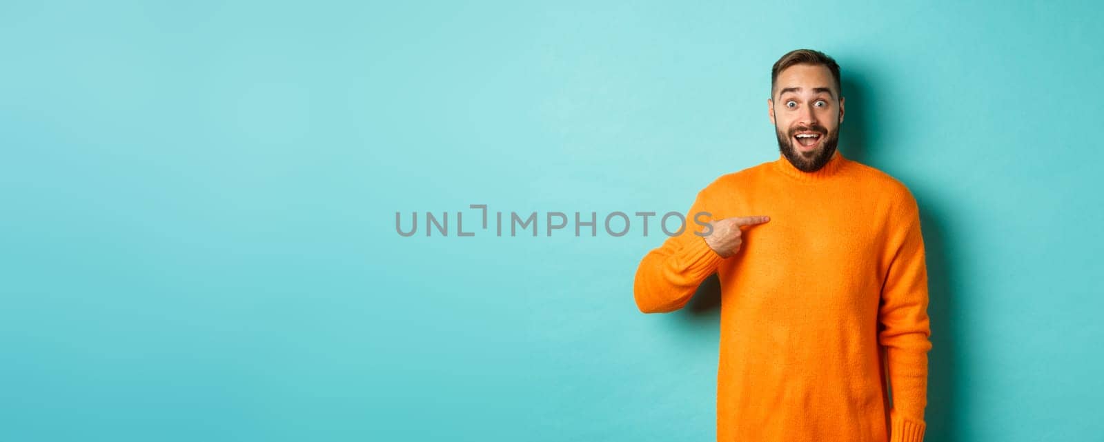 Excited man pointing at himself, looking amazed and happy, being chosen, standing over light blue background.