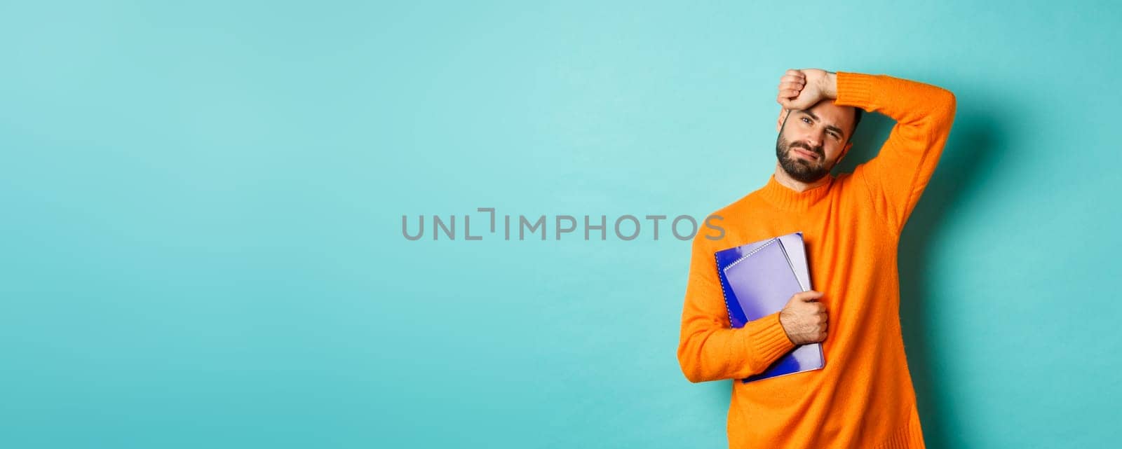 Education. Tired male student holding notebooks and wiping sweat off forehead, looking exhausted, standing in orange sweater against turquoise background.
