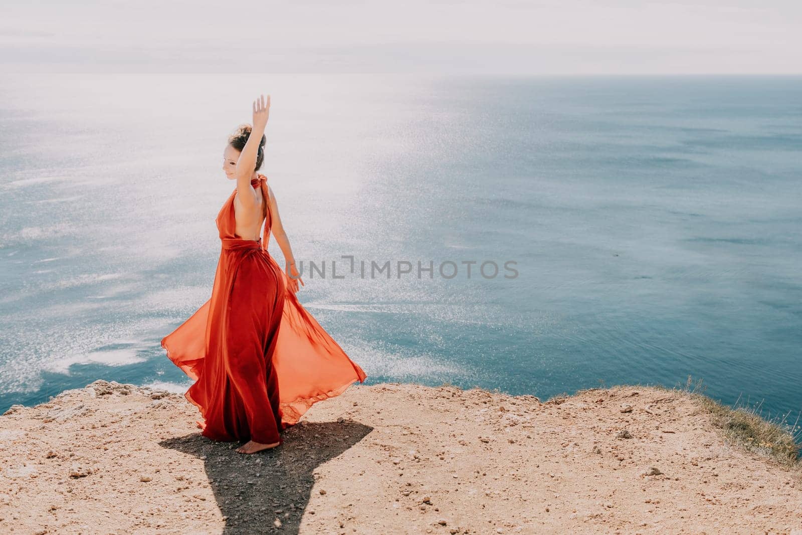 Woman in red dress on sea. Side view a Young beautiful sensual woman in a red long dress posing on a rock high above the sea on sunset. Girl on the nature on blue sky background. Fashion photo. by panophotograph