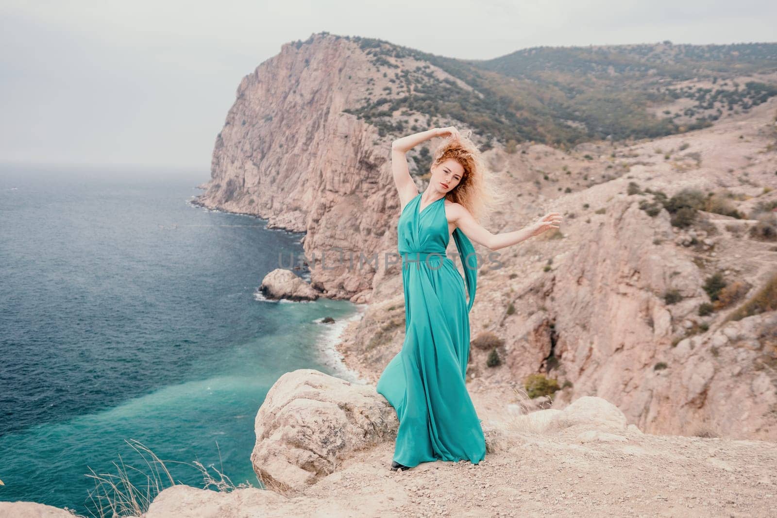 Side view a Young beautiful sensual woman in a mint long dress posing on a volcanic rock high above the sea during sunset. Girl on the nature on overcast sky background. Fashion photo