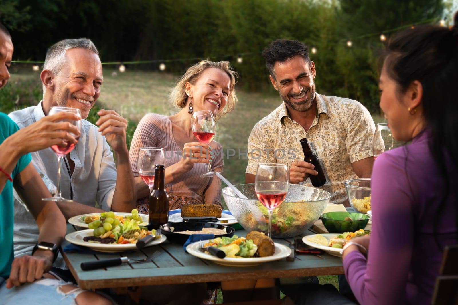 Friends laughing, talking and drinking wine at garden dinner party in the evening. by Hoverstock