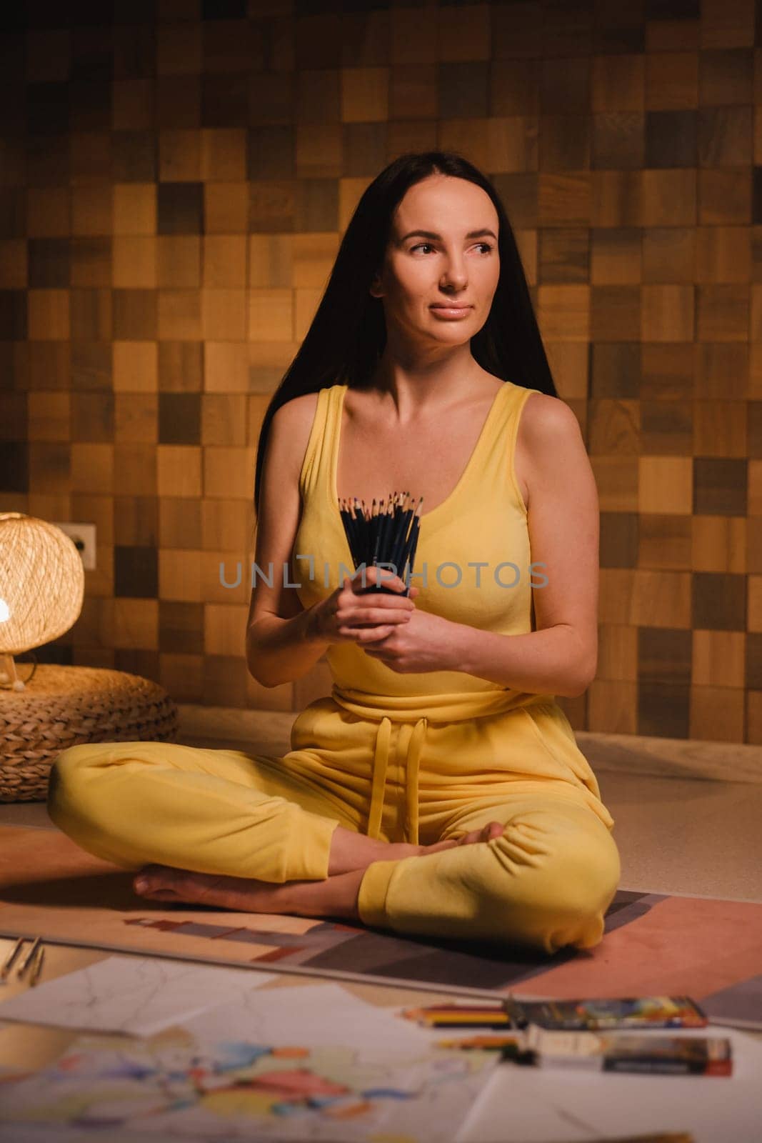 A girl in yellow clothes sitting on the floor holding pencils for drawing.