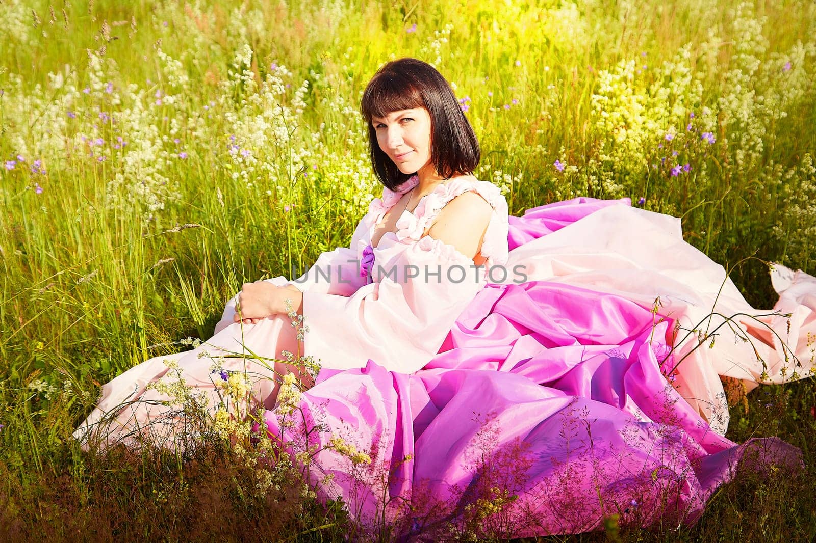 Beautiful girl in a lush pink ball gown in green field during blooming of flowers and blue sky on background. Model posing on nature landscape as princess from fary tale by keleny