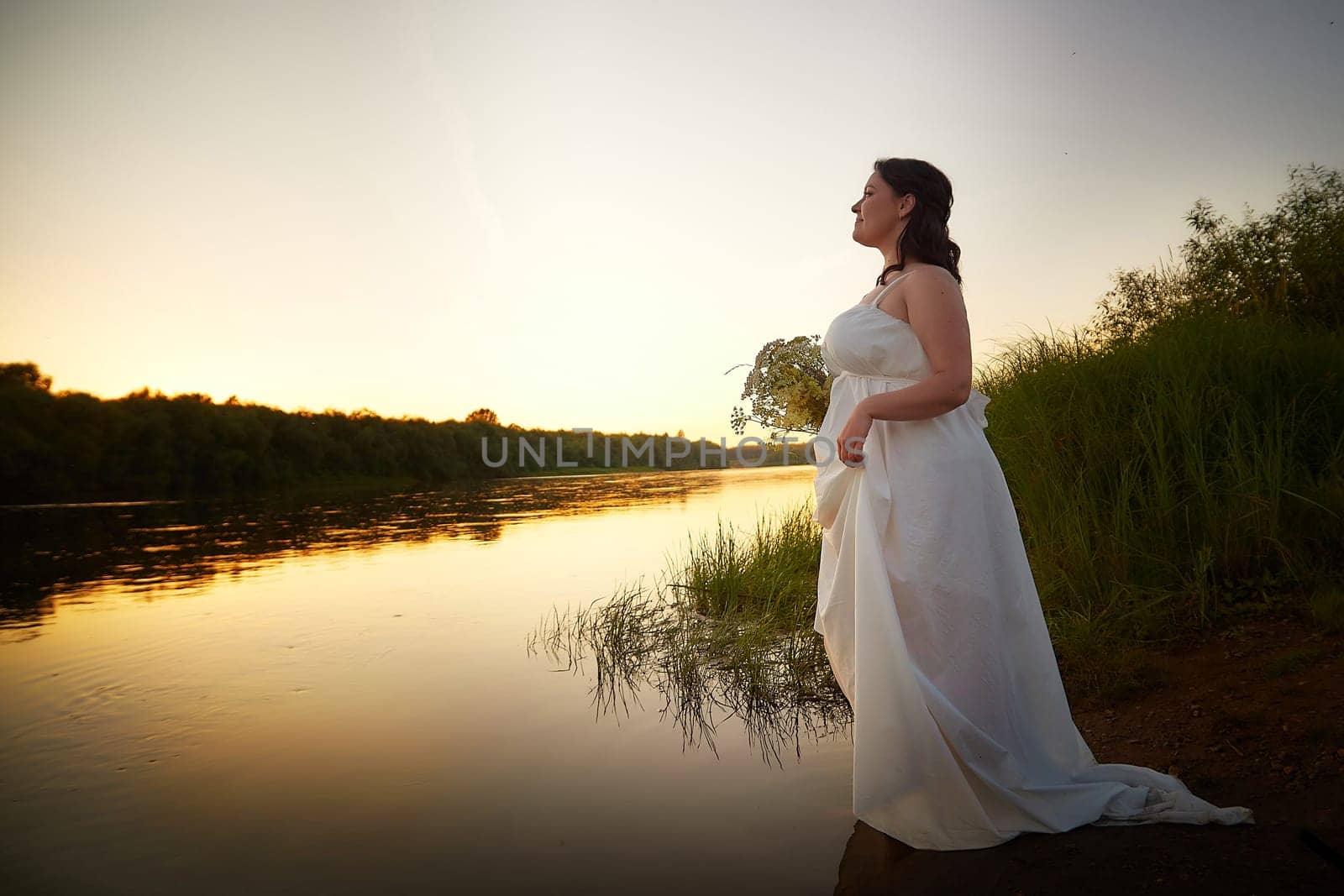 Slavic plump plump chubby girl in long white dress on the feast of Ivan Kupala with flowers and water in a river or lake on a summer evening by keleny