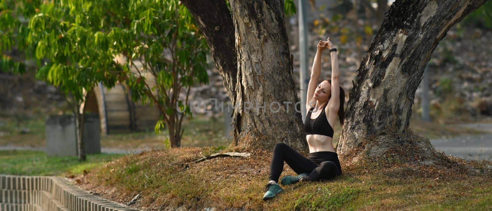 Horizontal full length view of athletic caucasian woman stretching arms, resting after workout on grass in summer park.