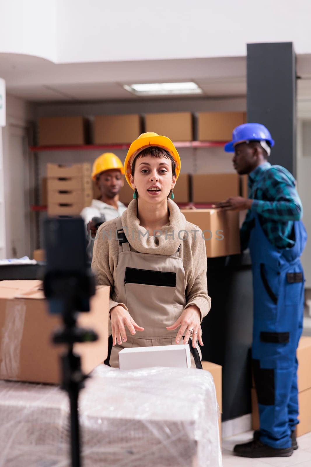 Warehouse operator showing buyer order picking and packing at smartphone camera. Young woman wearing protective helmet preparing parcel for dispatching and looking at mobile phone on tripod