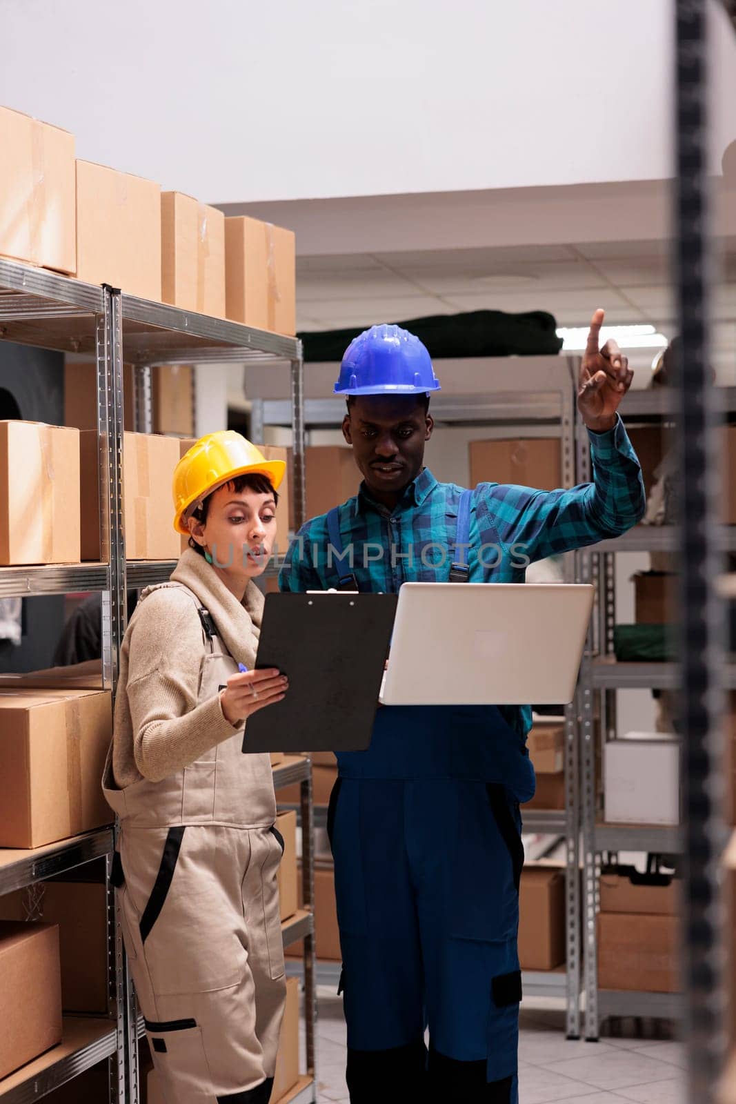 Warehouse employees comparing inventory data on clipboard and laptop. Diverse african american and caucasian coworkers wearing protective helmets checking parcels maintaining in storehouse