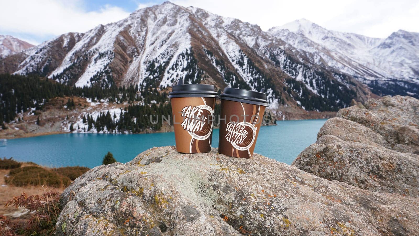 Paper cups with coffee on the background of a mountain lake. The color of the water is blue. A green forest grows on the hills. The high peaks are covered with snow. The cups are on a stone. Romance