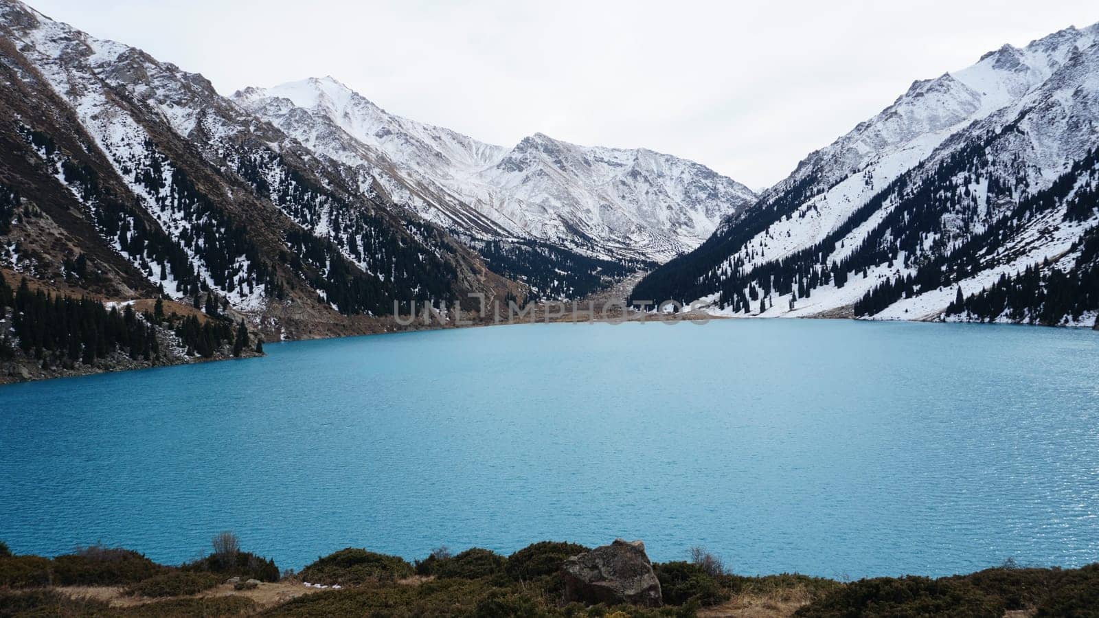 A mountain lake with blue water in winter. The sun's rays are reflected from the water. Green forest and grass grow on the hills. There are stones lying. The high peaks are covered with snow. Almaty