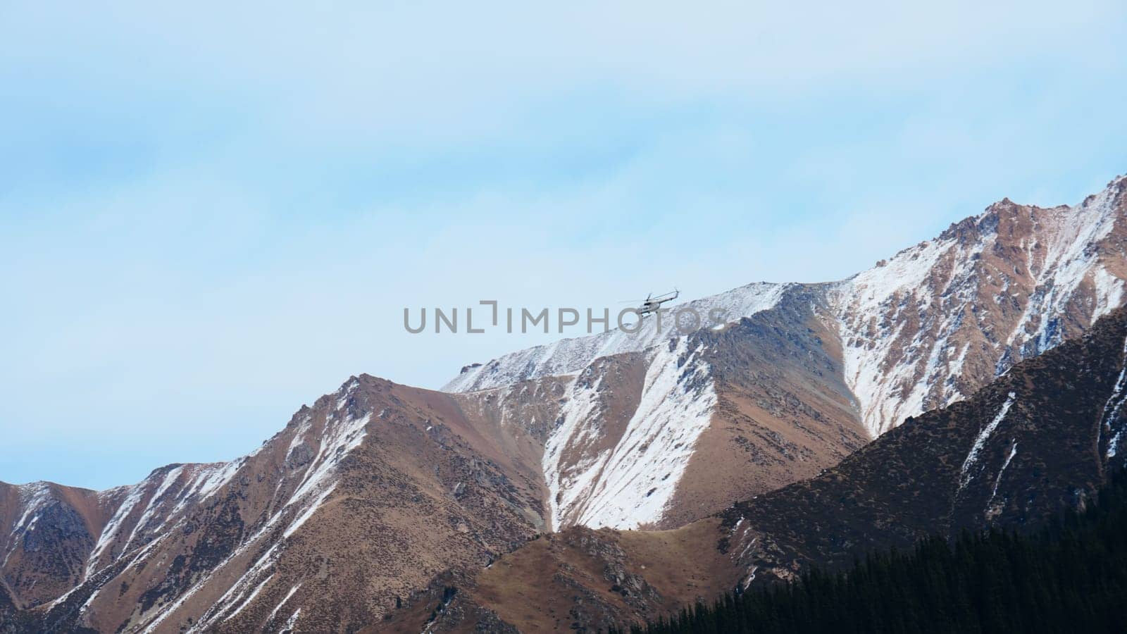 The helicopter is flying over the snowy mountains. Dark forest below, yellow grass on the hills. Steep cliffs and large rocks. Blue sky in clouds. Rescue helicopter. Almaty Mountains, Kazakhstan